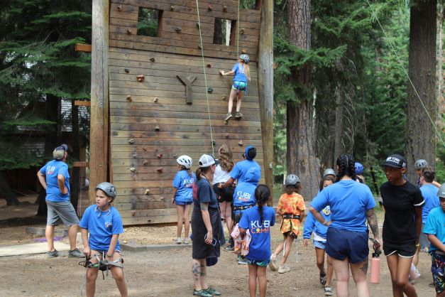 A camper participates in rock climbing while other campers look on.