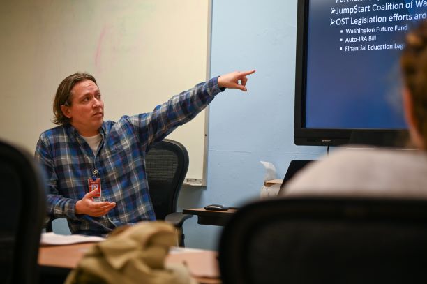 : Man at the front of the room, addressing others in the room.